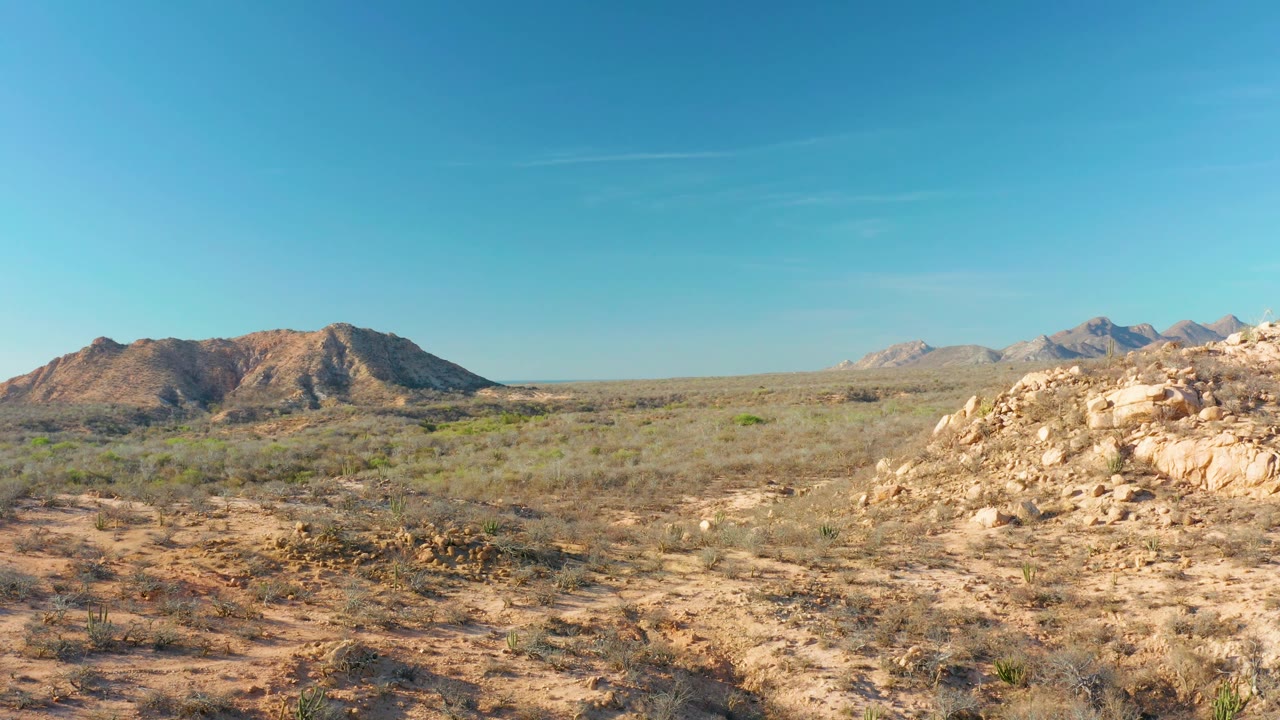 At 0:01, the Cabo Pulmo desert remains in a wide view, with low foreground plants, tan rock on the right, distant ridges, and no visible people.