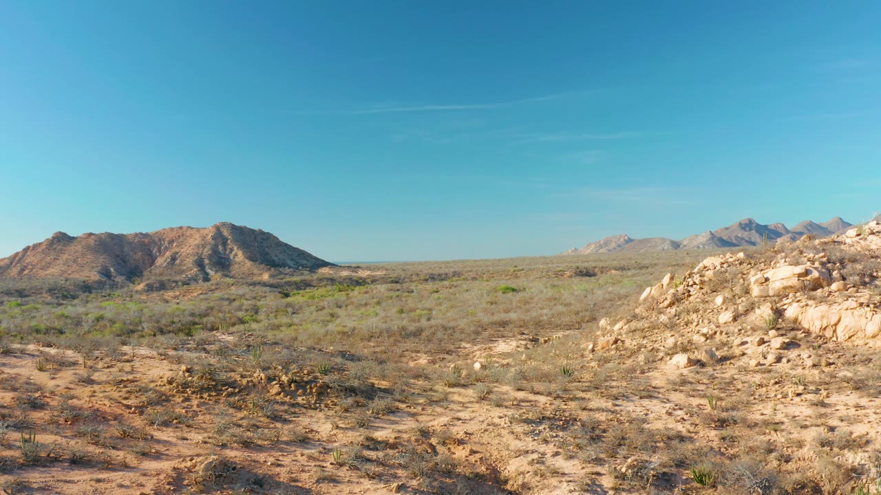 At 0:02, sparse desert vegetation stretches across the Cabo Pulmo foreground, with a sunlit rocky rise on the right and distant mountains under clear sky, no visible people.
