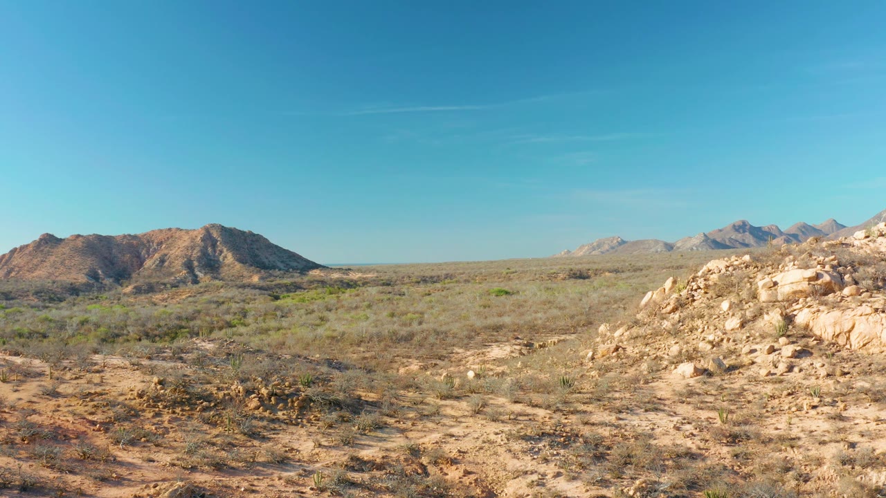 At 0:03, the Cabo Pulmo landscape shows cactus clusters and sandy ground in front of tan rocks, with low hills and sharper distant mountains, no visible people.