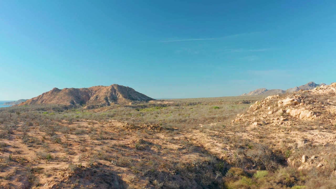 Cabo Pulmo arid scrubland fills the foreground with a low rocky hill left of center, pale mountains in the distance, a bright rock outcrop on the right, and no visible people.
