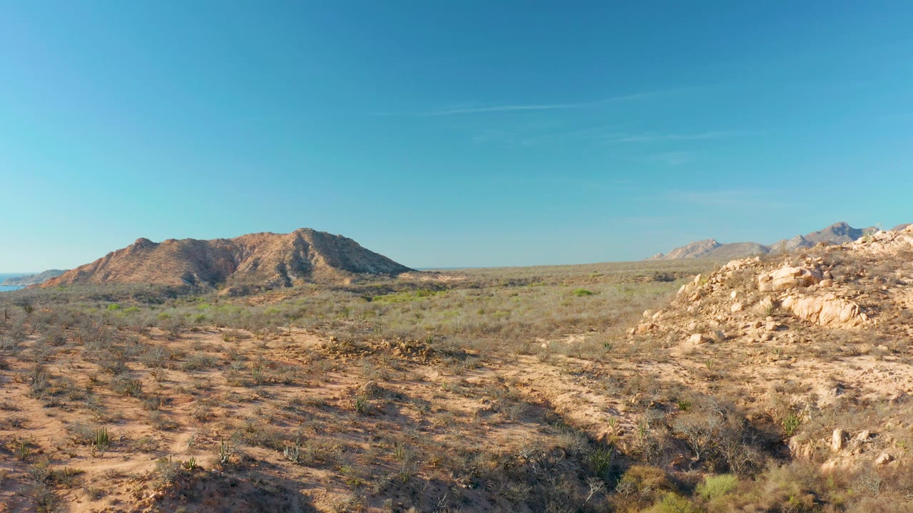 The Cabo Pulmo desert plain appears sunlit and open, with textured sand and brush in the foreground, brown rocky hills in back, distant mountains to the right, and no visible people.
