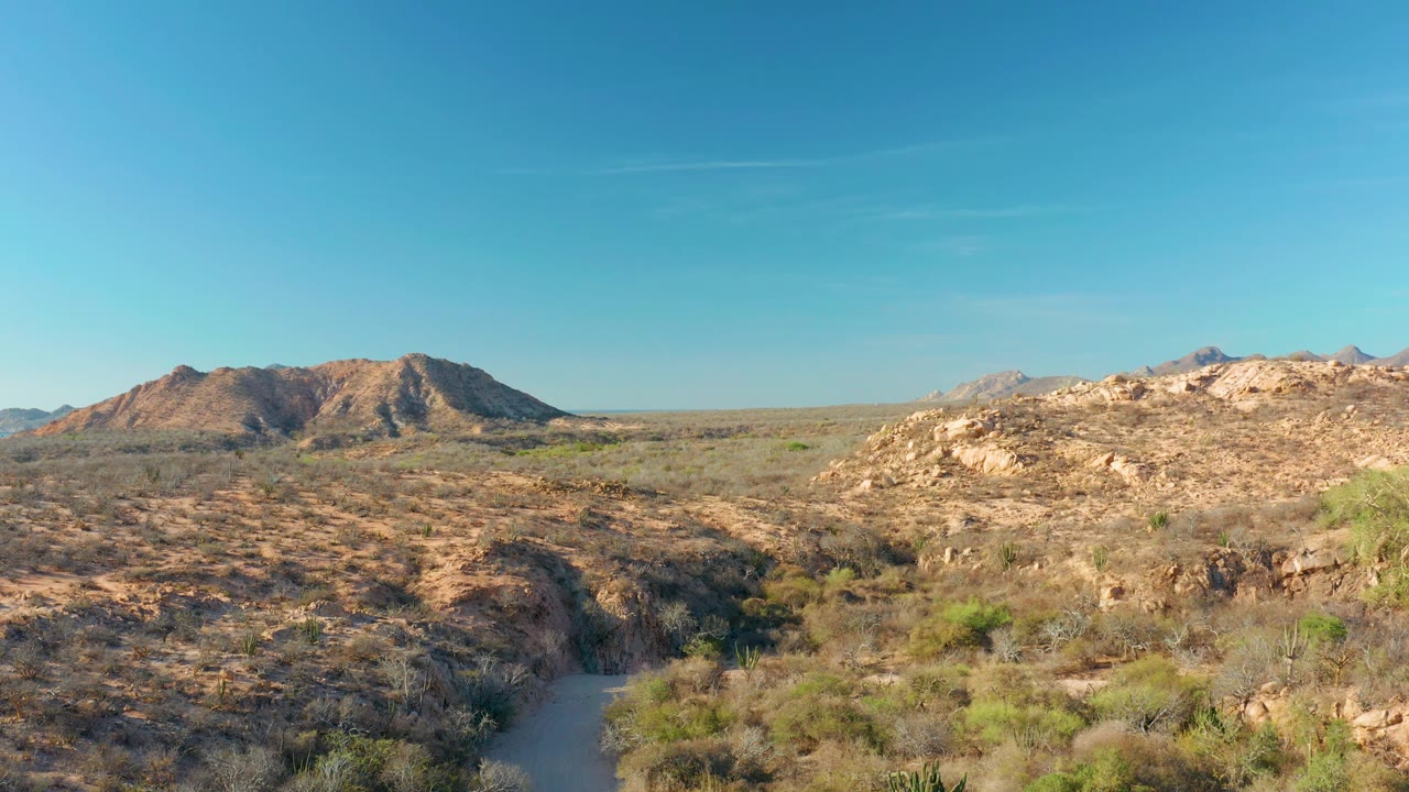 Mid-clip view of the rocky desert wash and scrub near Cabo Pulmo, with brown hills, distant mountains, blue sky, and no visible people.