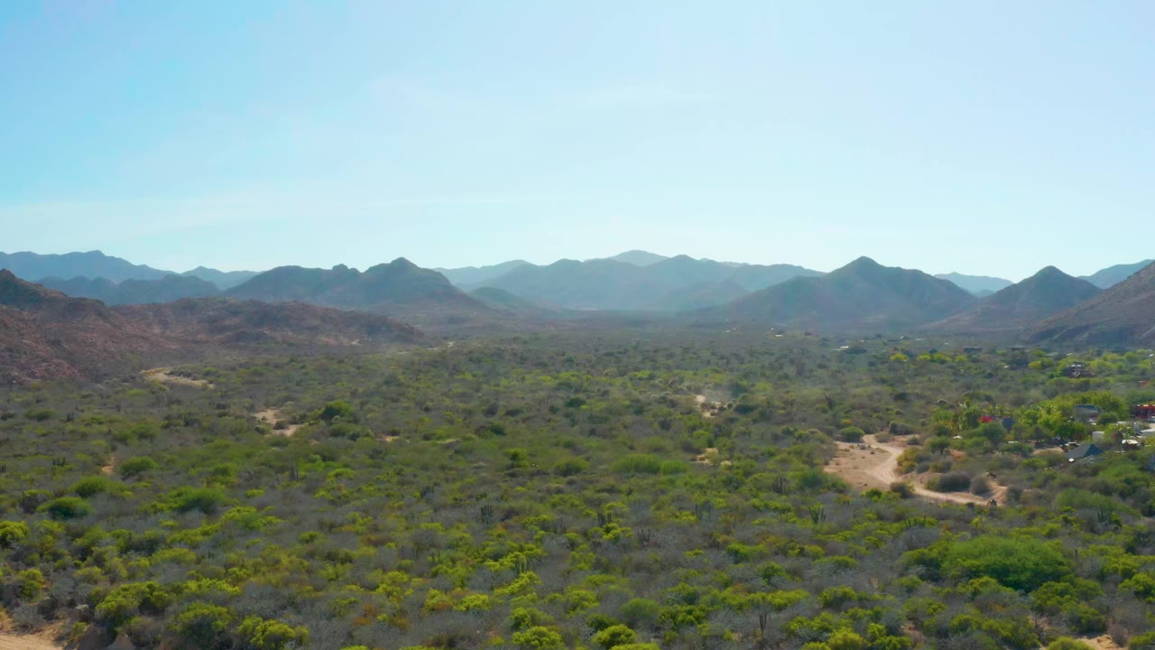 Opening aerial frame over Cabo Pulmo desert valley with a pale dirt road through green-gray brush, layered mountains on the horizon, clear blue sky, and no visible people.