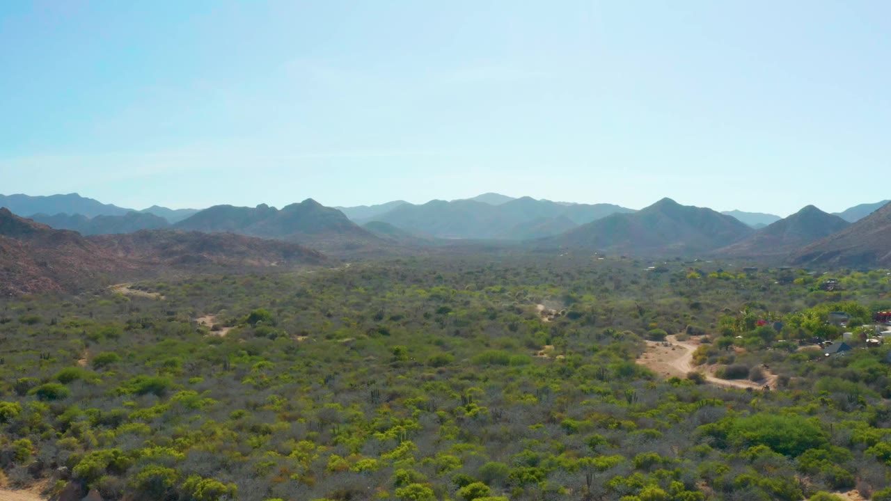 Aerial desert valley with winding road, brush, mountains, and a faint dust trail.