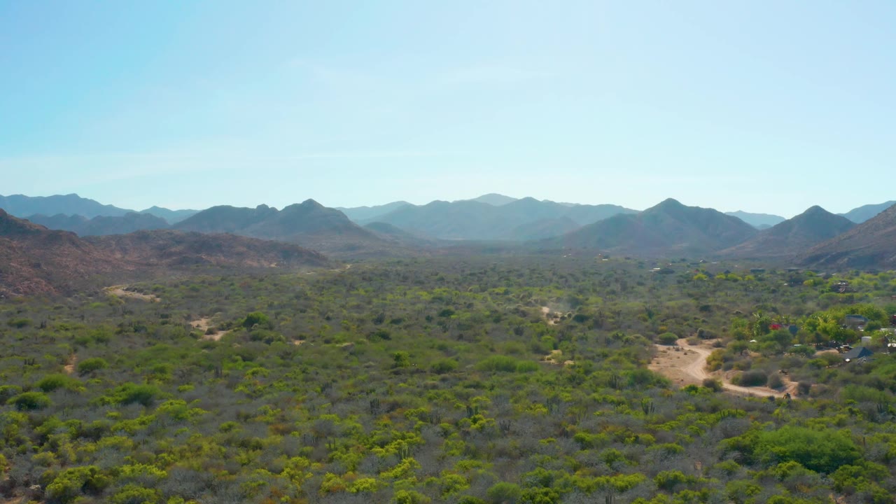 High aerial frame in Cabo Pulmo showing a dusty road area among sparse trees and brush, with reddish foothills and hazy mountain layers under a clear sky, no visible people.