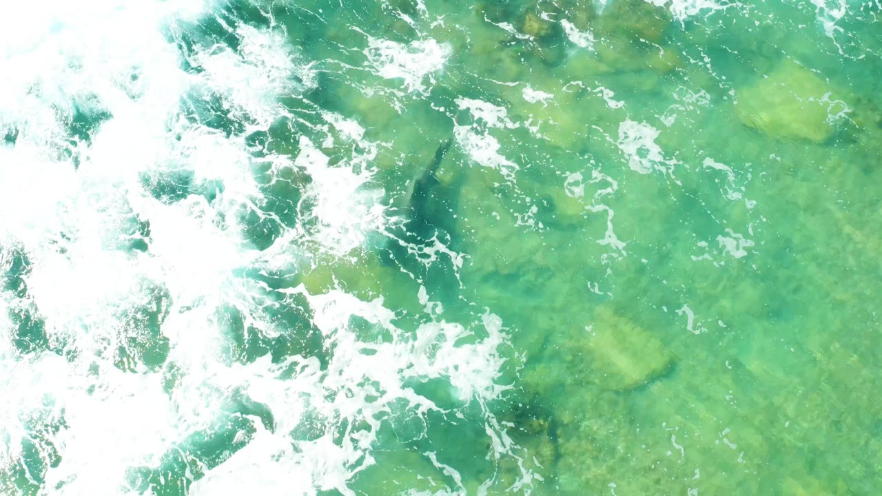 Overhead Cabo Pulmo frame with dense white foam curling across the left side and green-blue water revealing rocks beneath, no visible people.