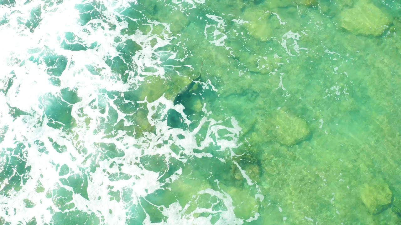 Overhead frame from Cabo Pulmo with a bright white foam band across the left and center over pale green turquoise water and visible rocks, no visible people.