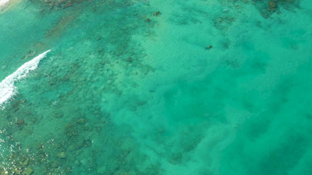 Aerial Cabo Pulmo frame showing clear turquoise water, darker reef patches under the surface, and a narrow white foam streak along the left side, no visible people.