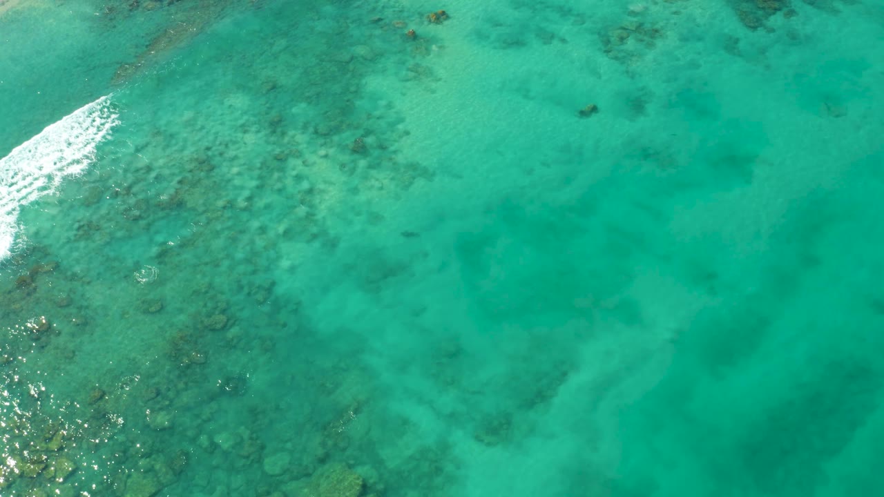 Aerial view over Cabo Pulmo shallow water with coral and rock textures below the surface and a diagonal white foam edge entering from the left, no visible people.