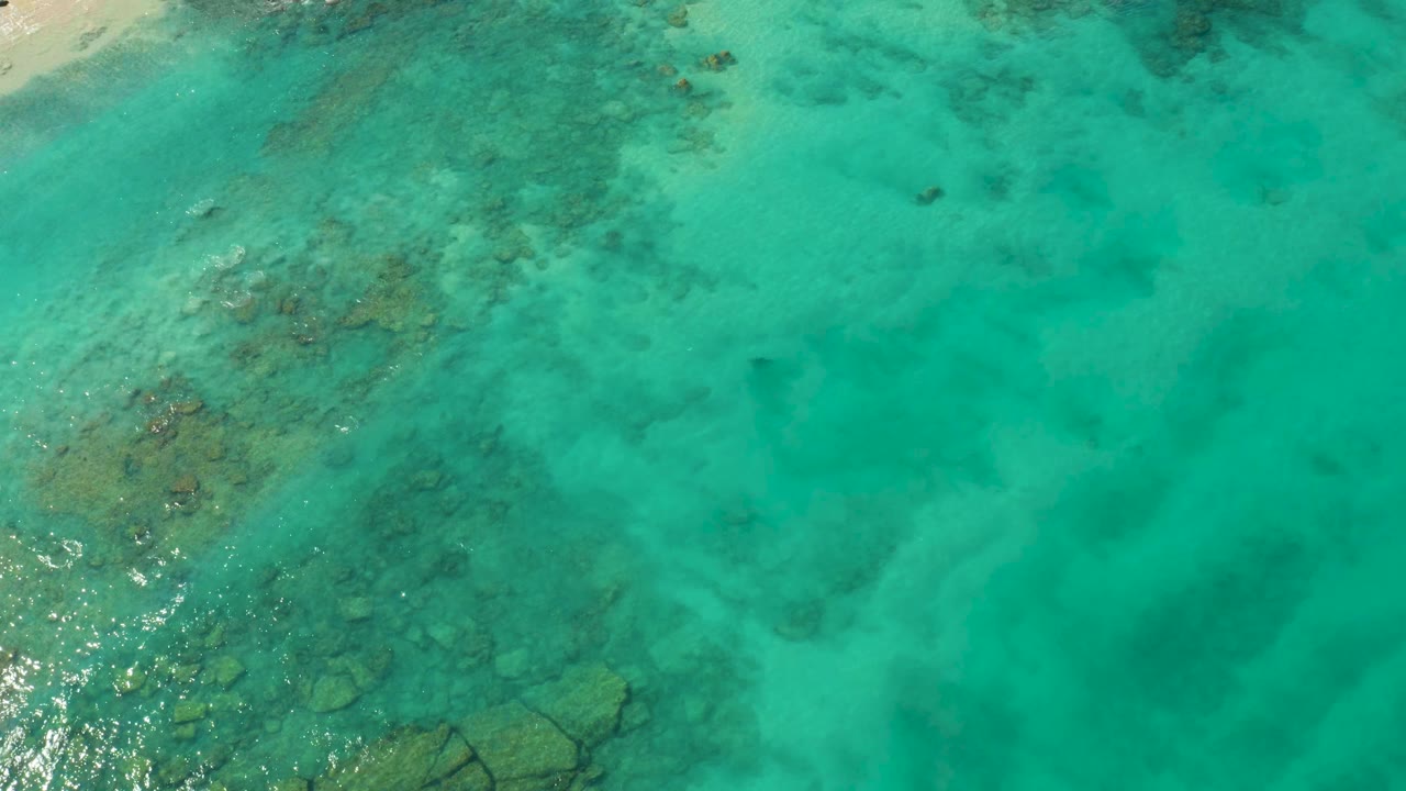 Overhead view of clear turquoise ocean in Cabo Pulmo, Mexico, with submerged rocks and reef patches visible under the water and no visible people.