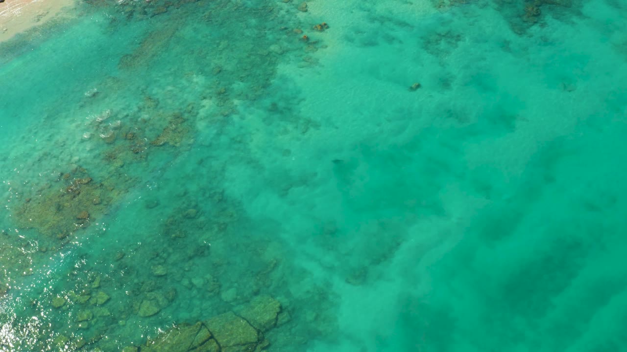 Aerial frame over Cabo Pulmo's blue-green water, with reef patches clustered toward the left side and open turquoise water to the right, no visible people.
