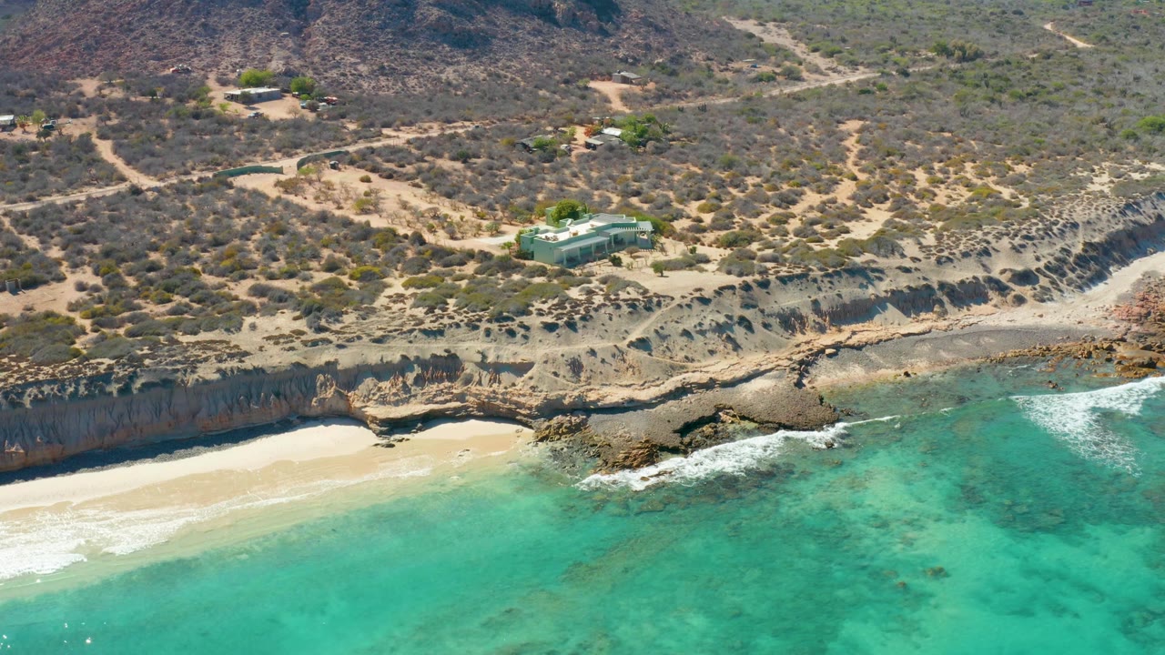 Turquoise ocean reaches a pale sandy beach beneath eroded rocky cliffs and scrub terrain.