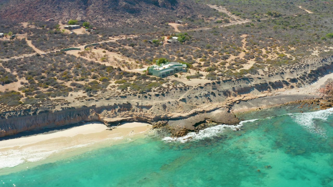 Cabo Pulmo aerial frame showing more white foam around the rocky point, a sandy cove at left, turquoise shallows at right, dry hillside tracks, small buildings, and no visible people.