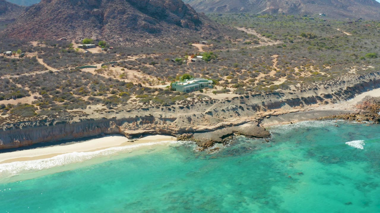 Opening aerial frame over Cabo Pulmo showing turquoise water, pale sand, rocky cliff edges, dry hillside vegetation, dirt tracks, distant mountains, a small hillside structure, and no visible people.