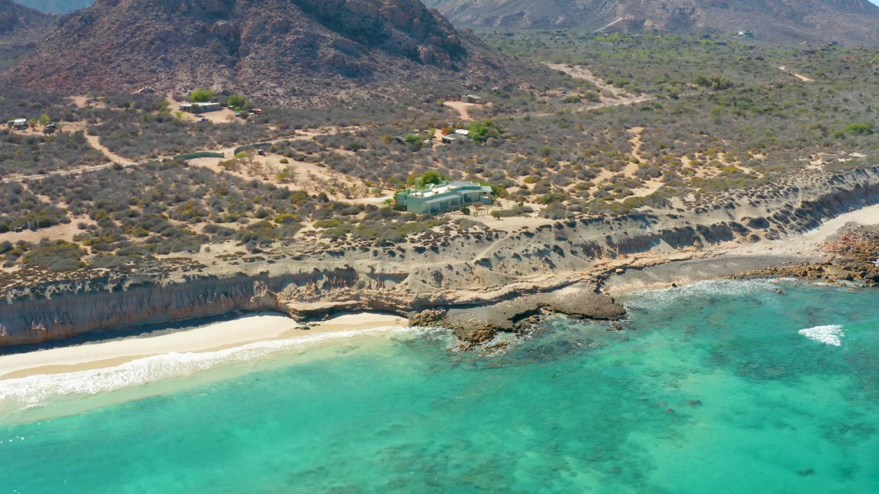 Aerial frame over Cabo Pulmo with clear turquoise water filling the foreground, a thin white surf line on the sand, rocky cliffs, dry scrubland, dirt tracks, distant hills, and no visible people.