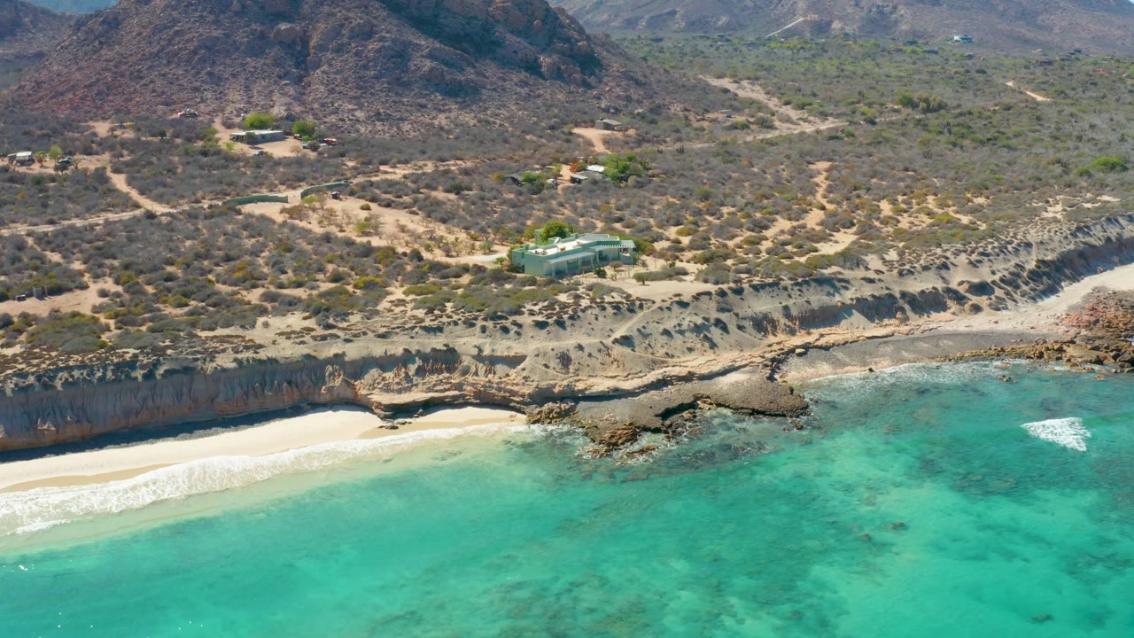 Cabo Pulmo aerial view showing bright turquoise water in the foreground, sand along the lower cliff, rocky outcrops near shore, sparse dry vegetation above, dirt roads, a small structure, and no visible people.