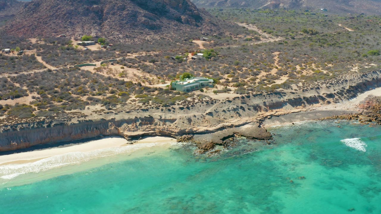 Oblique drone view of a sandy beach under cliffs with turquoise water and dry hills inland.