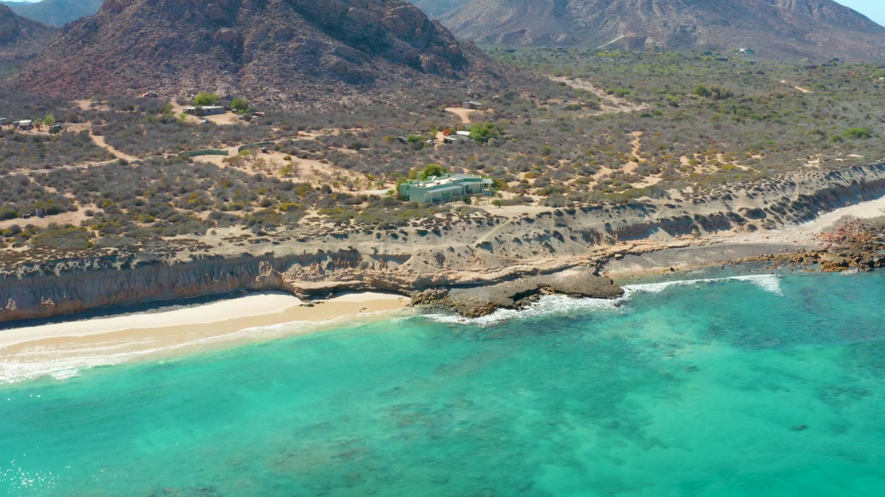 Aerial view at Cabo Pulmo with clear turquoise water in the foreground, a pale sandy beach to the left, rocky shoreline across the middle, dry hillside terrain, and no visible people.