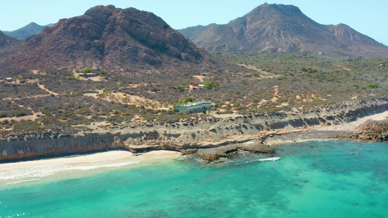 Drone view of clear blue-green water beside a pale beach and rocky coastal bluffs.