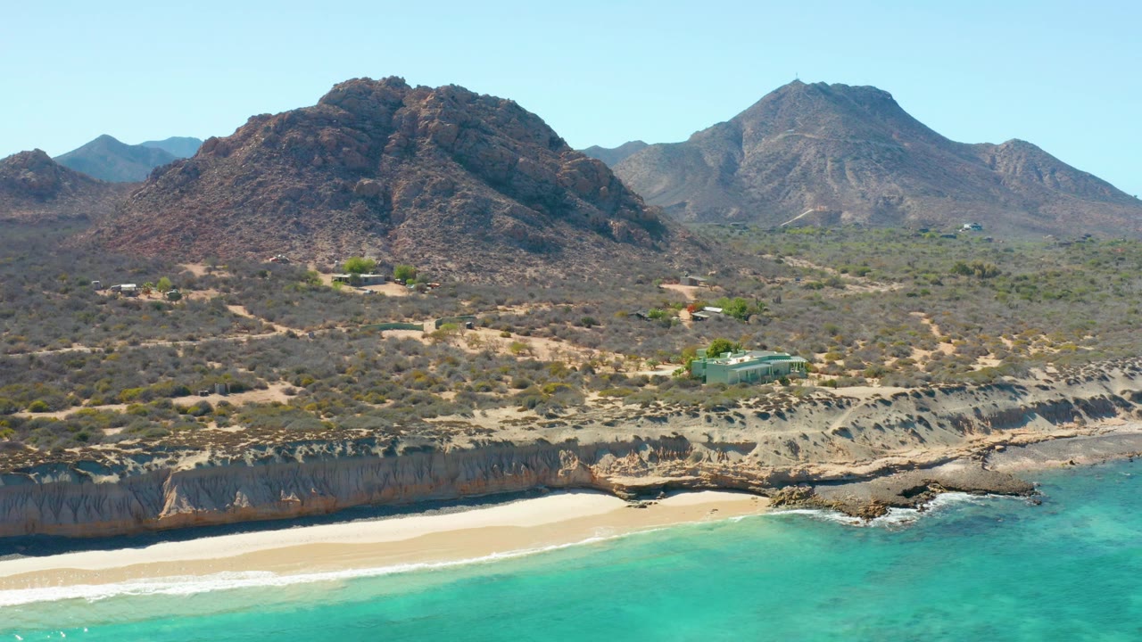 Opening aerial frame over Cabo Pulmo with turquoise water at the bottom, a pale sand beach below eroded cliffs, scrub-covered land, small structures, and mountains beyond, with no visible people.
