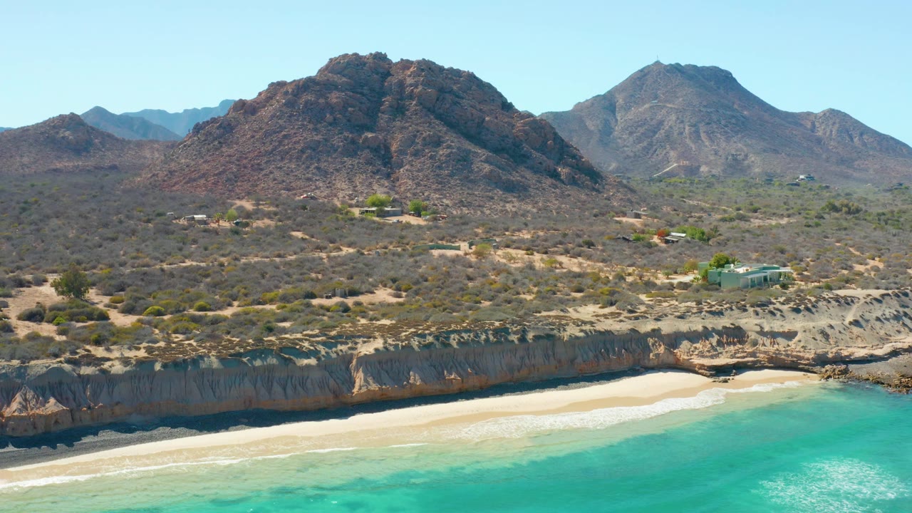 Aerial view in Cabo Pulmo, Mexico, with turquoise water below a pale sandy beach, eroded cliffs, arid scrubland, mountains, scattered small buildings, and no visible people.