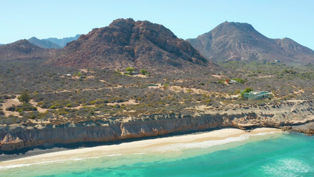 Wide drone view of a sandy cove, turquoise shoreline, rocky ledge, and dry mountains.
