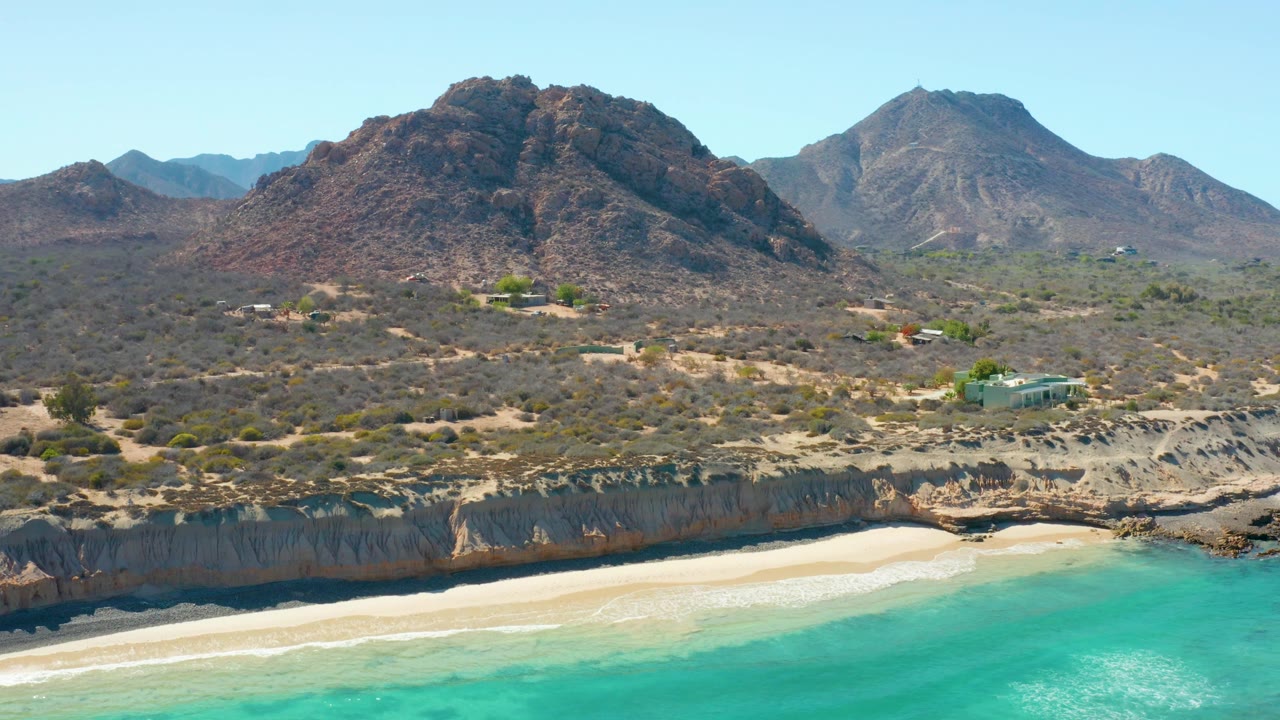 Aerial frame from Cabo Pulmo, Mexico, with bright turquoise water at the lower edge, a clean band of sand, dark rocky cliffs, scrubby desert land, rugged mountains, and no visible people.