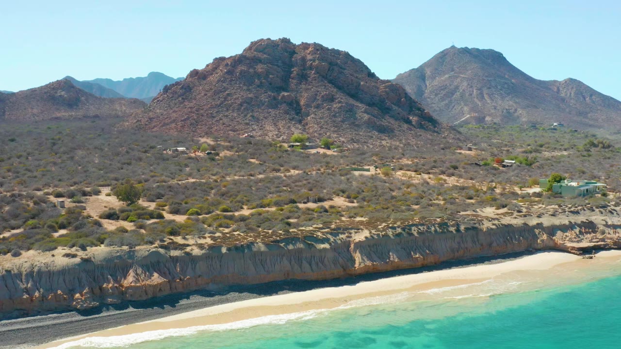 Aerial frame at Cabo Pulmo showing a longer stretch of sandy shoreline, clear turquoise water, cliff shadows, low desert vegetation, small inland buildings, rocky hills, and no visible people.