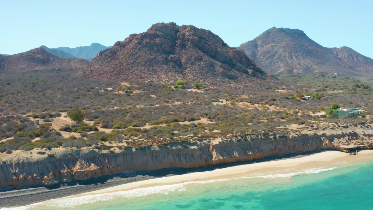 Coastal aerial view with waves, sandy beach, cliff band, desert shrubs, and mountains.