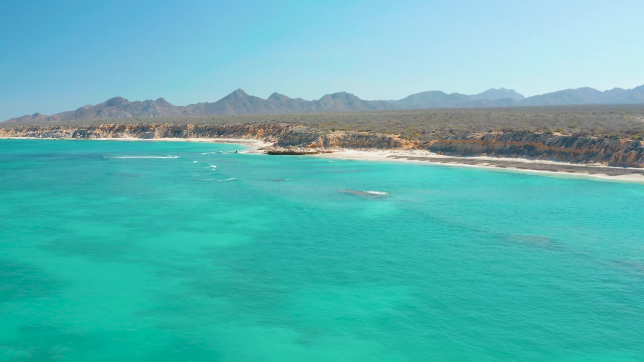 Cabo Pulmo coastline seen from above with broad turquoise water, a sandy shoreline, darker reef or rock patches under the water, distant mountains, and no visible people.