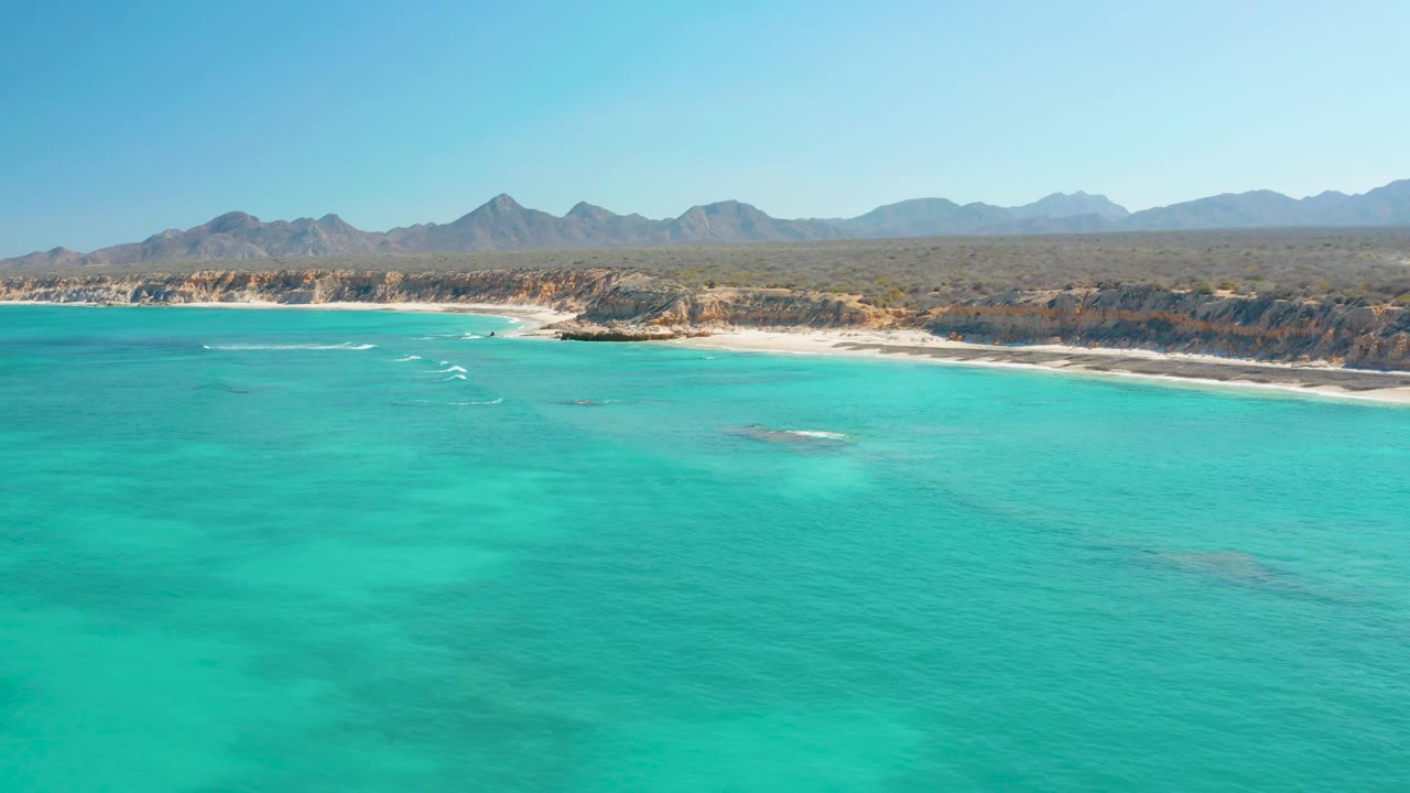 Aerial Cabo Pulmo frame with turquoise ocean in the foreground, white wakes near the shore, a pale beach, low cliffs, mountains on the horizon, and no visible people.