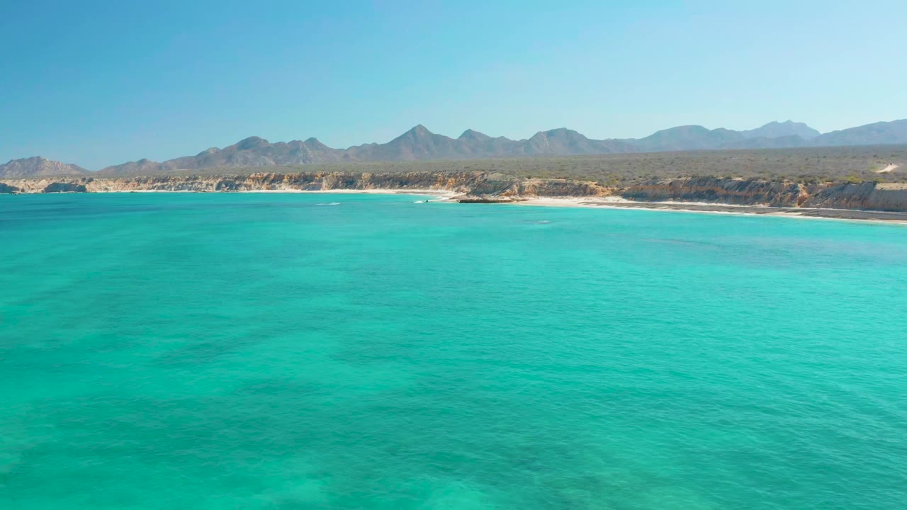 Wide aerial view of turquoise water facing the Cabo Pulmo shoreline, with a sandy beach, arid land, distant mountains, blue sky, and no visible people.