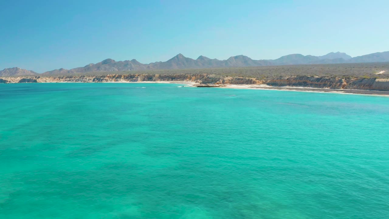 Aerial frame over clear blue-green ocean near Cabo Pulmo, showing pale surf along the beach, scrubby coastal land, distant mountains, and no visible people.
