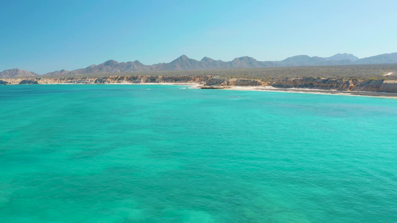 Drone-height view across turquoise sea toward the Cabo Pulmo beach, with subtle darker patches under the water, a long shore, mountain silhouettes, and no visible people.