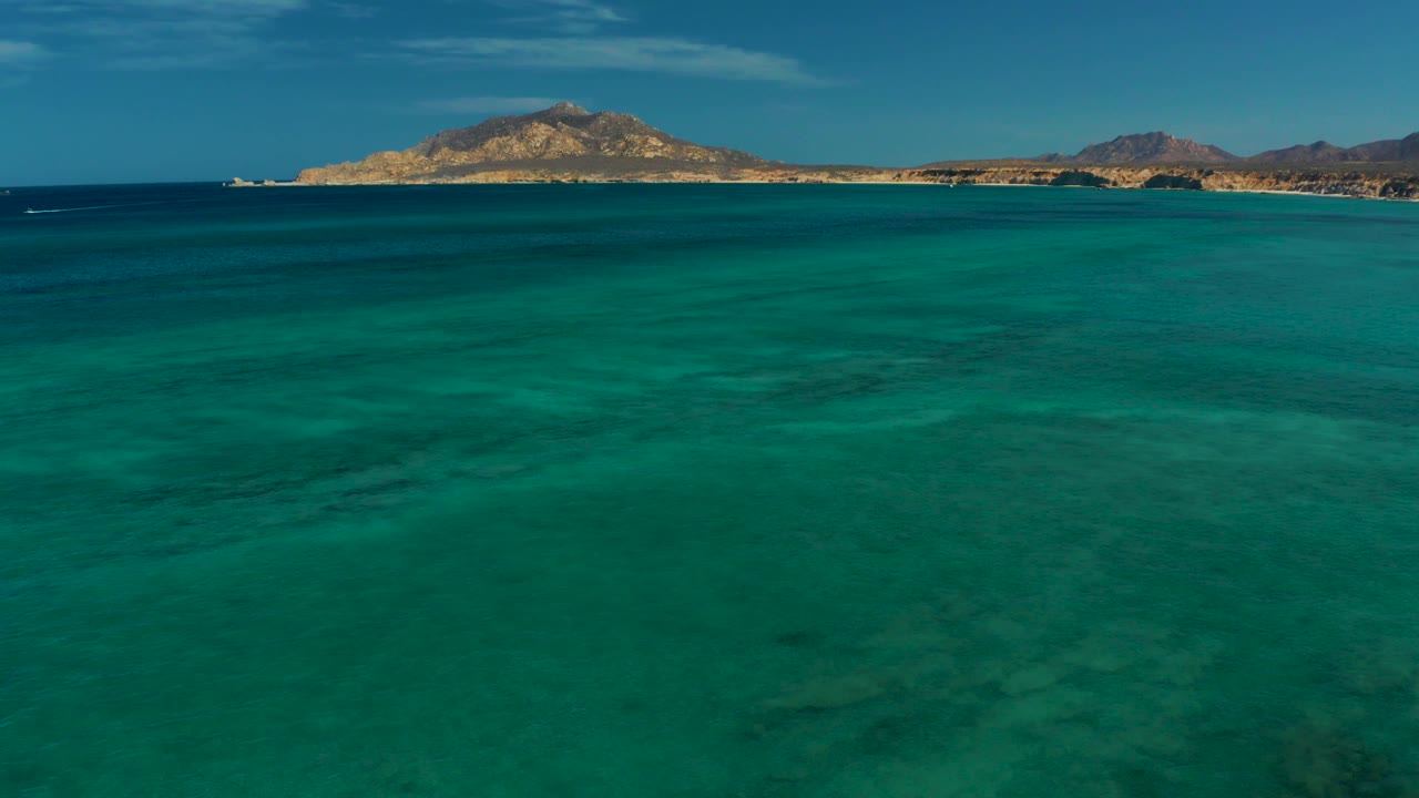 A broad band of shallow turquoise sea stretches toward the Cabo Pulmo shore, with rocky hills beyond and no visible people.