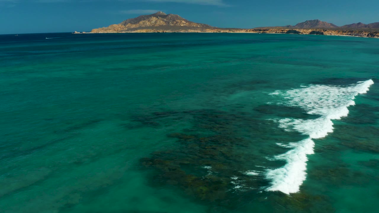 Aerial view in Cabo Pulmo, Baja California Sur, showing turquoise water, dark coral reef patches, white surf breaking near center frame, distant low hills, and no visible people.