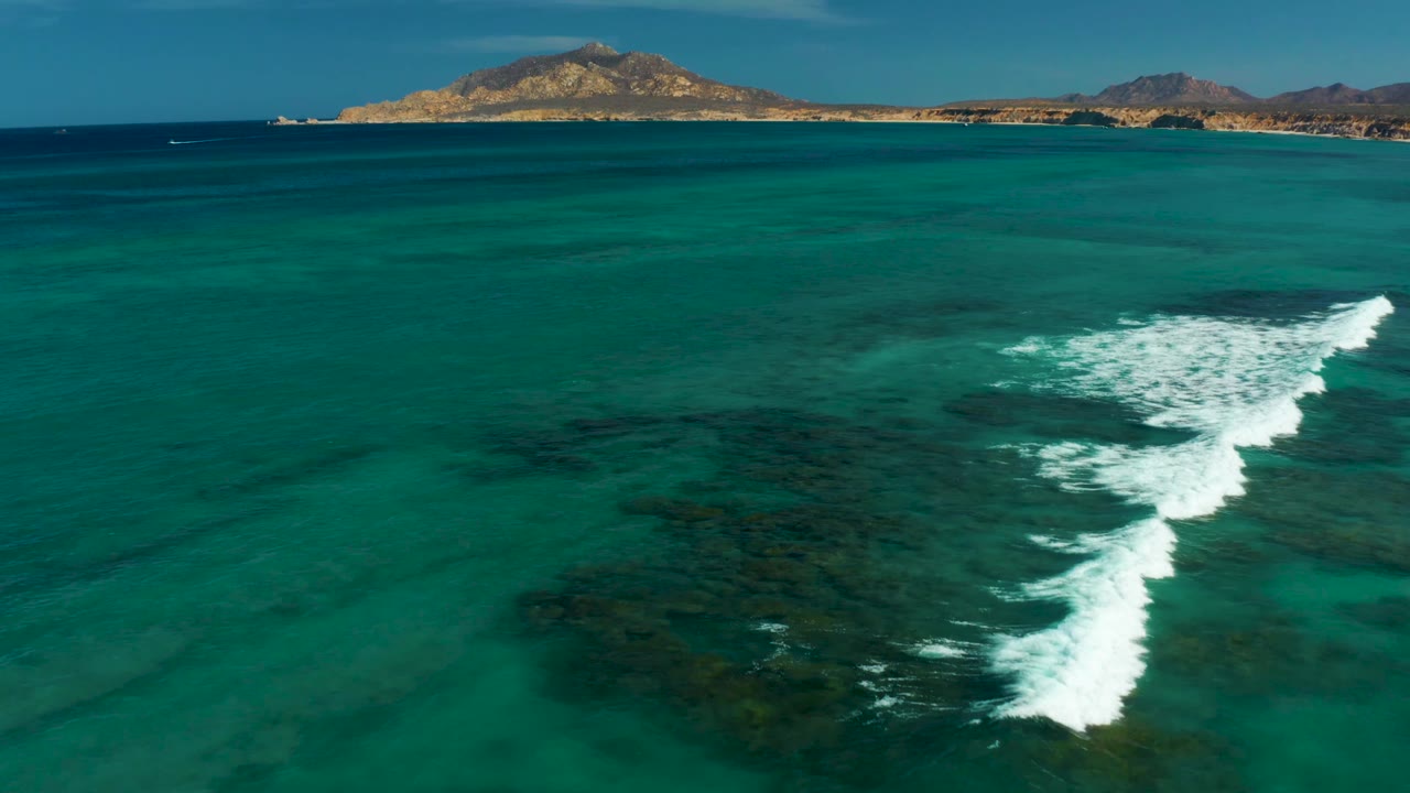 Aerial Cabo Pulmo frame with a longer white foam line crossing the reef, clear green-blue shallows below, low coastal hills along the horizon, and no visible people.