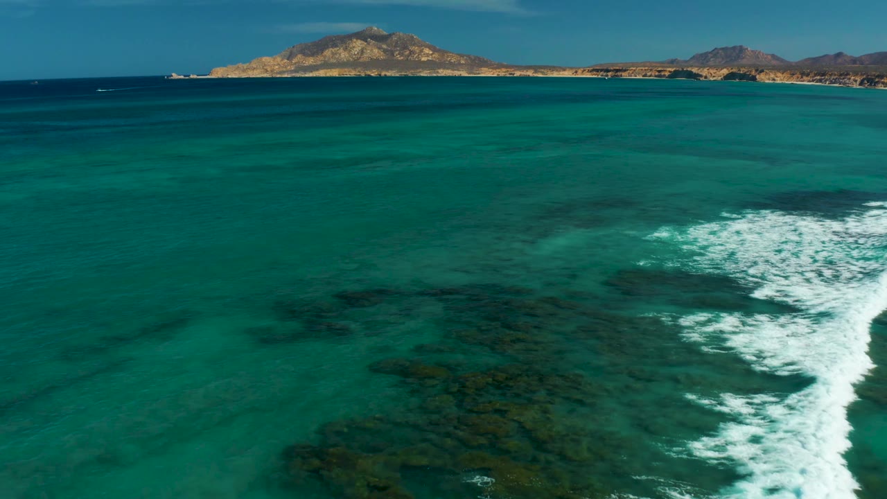 Aerial view over Cabo Pulmo shallows where white surf sits closer to the right side of frame, dark reef patches show through the water, hills line the horizon, and no visible people appear.