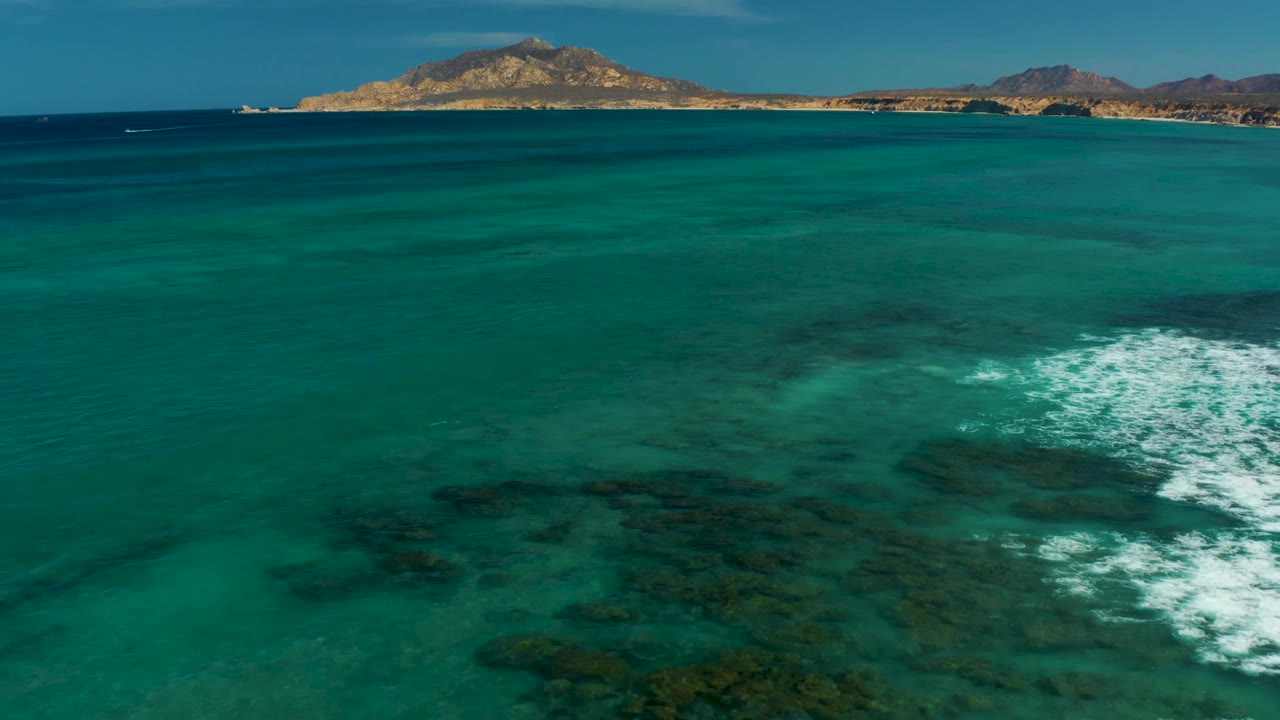 Aerial reef water with white foam near the lower right and a distant hilly coastline.