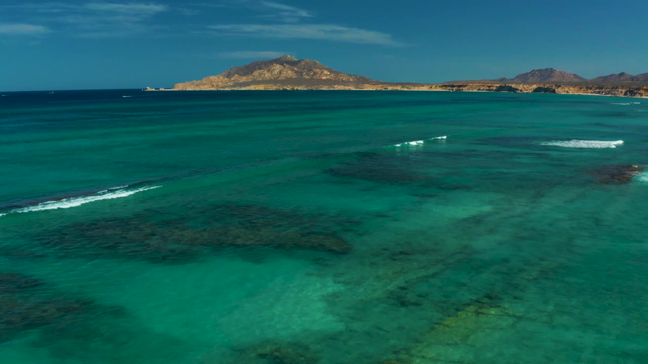 Aerial view over turquoise ocean water in Cabo Pulmo, Baja California Sur, with dark reef patches below the surface, a thin line of breaking foam, distant dry hills, and no visible people.