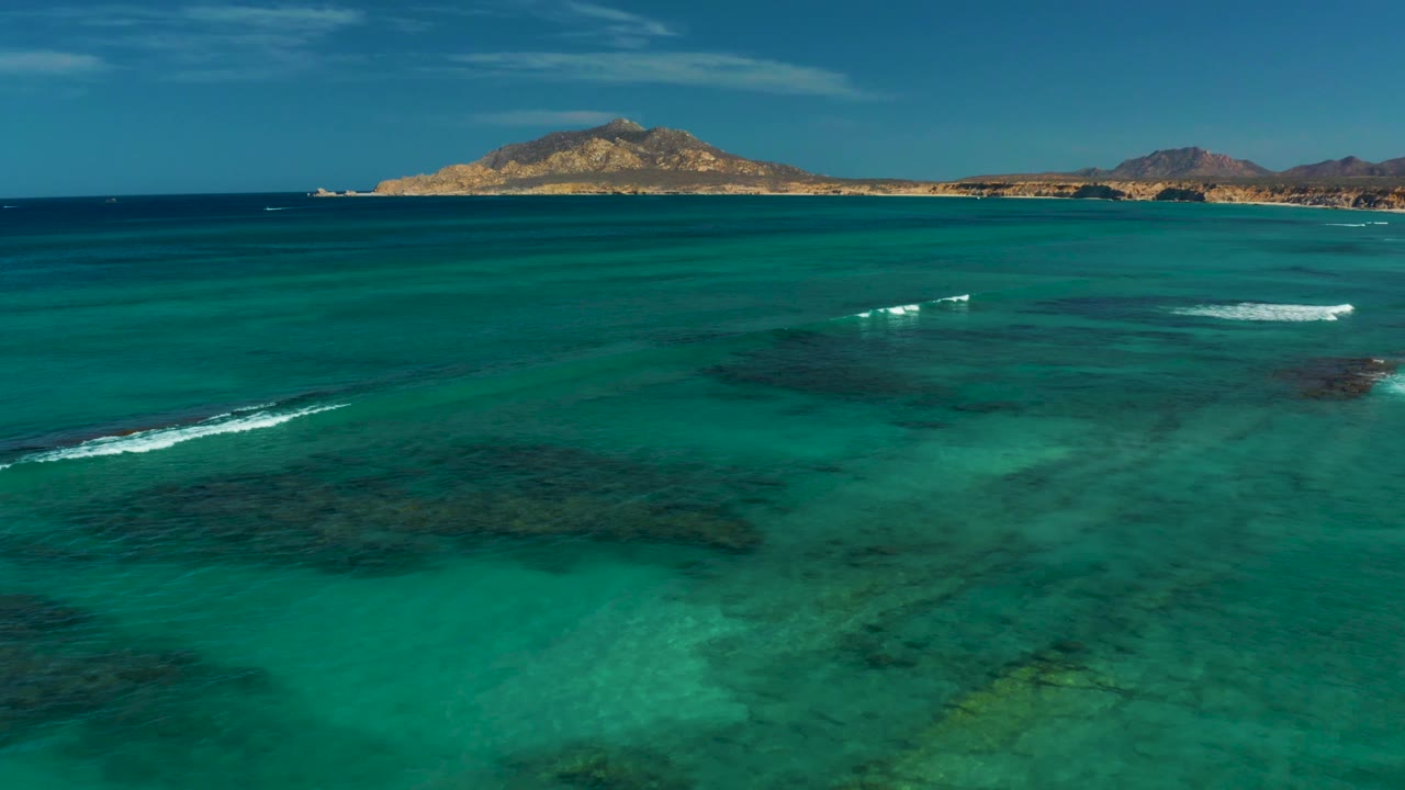 Aerial Cabo Pulmo frame with a white foam line crossing shallow turquoise water, darker underwater reef areas in the lower half, distant hills across the horizon, and no visible people.