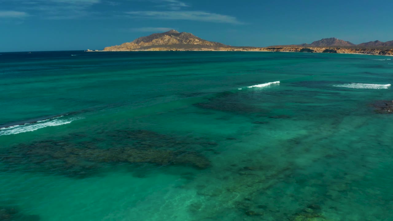 Aerial view of Cabo Pulmo water where white surf breaks over a dark reef patch, with broad turquoise shallows in front, a low tan coastline and hills in back, and no visible people.