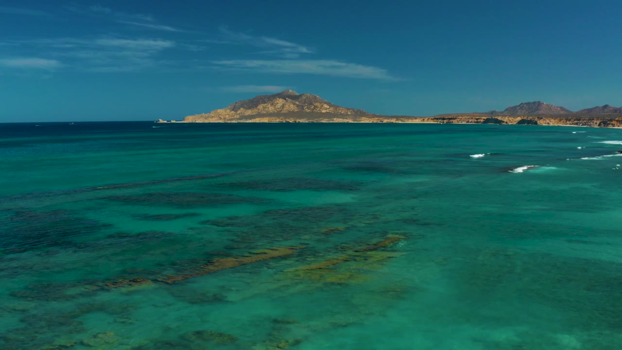 At 0:00, turquoise shallow water and darker seabed patches lead toward a distant island coastline in Cabo Pulmo, Mexico, with small whitecaps and no visible people.
