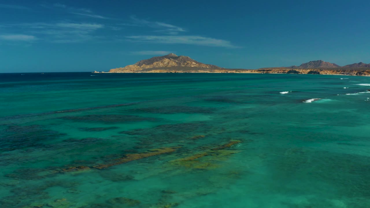At 0:01, the aerial composition holds over transparent turquoise sea with submerged reef shapes, a low island across the horizon, and no visible people.