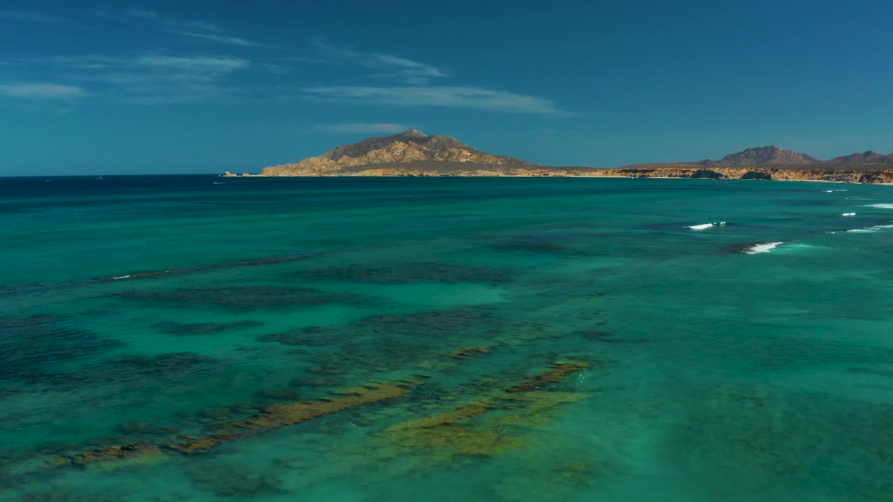 At 0:02, clear Cabo Pulmo water fills the frame with green and teal reef patches below, while a rocky island and low mountains sit along the top edge with no visible people.