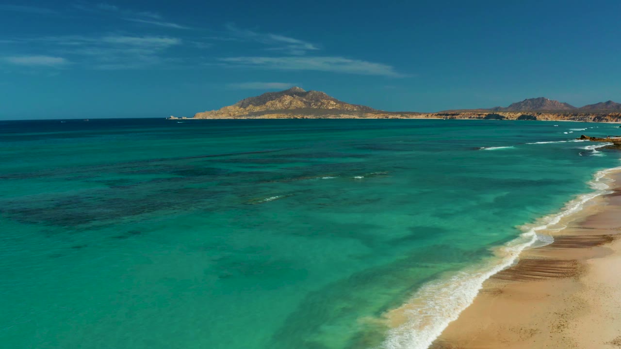 Aerial coastal frame in Cabo Pulmo showing clear shallow water, darker underwater patches, a beach running along the right edge, and no visible people.