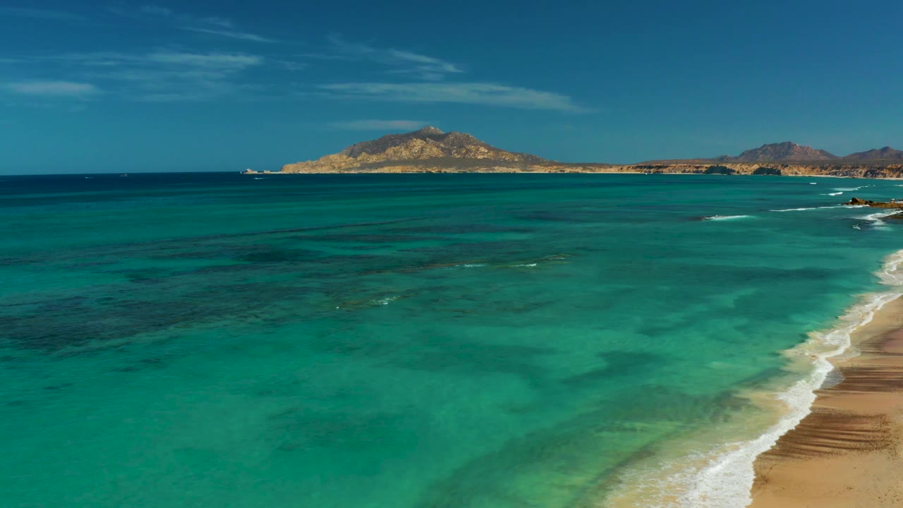 Cabo Pulmo shoreline frame with bright turquoise shallows in the foreground, a diagonal white wave line at right, a distant island centered on the horizon, and no visible people.