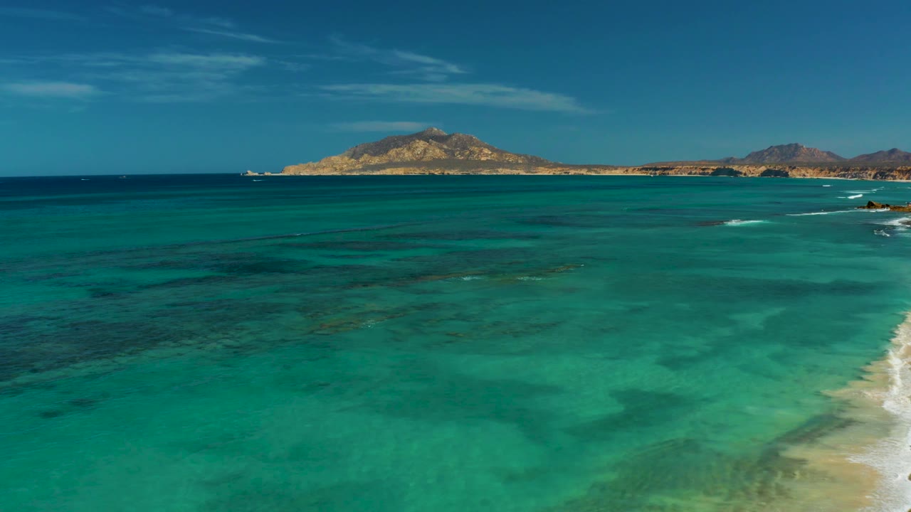 Mid-clip aerial view across open turquoise water in Cabo Pulmo, with the sandy beach clipped along the right side, low surf marks, distant landforms, and no visible people.