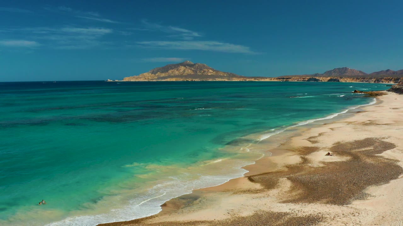 Aerial view in Cabo Pulmo, Mexico, showing turquoise water washing onto pale sand with distant hills under a clear blue sky and a tiny swimmer near the lower left.