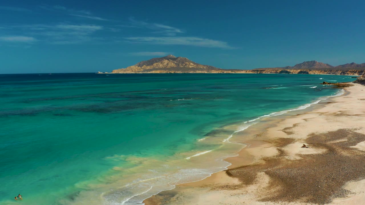 Cabo Pulmo aerial view with the shoreline running diagonally across the frame, turquoise water on the left, sand on the right, distant hills, and tiny people near the lower left edge.